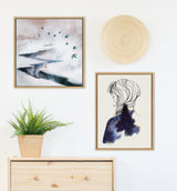 Interior wall with two framed artworks: abstract landscape with flying birds in muted tones and line-drawn female profile filled with a dark starry sky, above light wood dresser with leafy plant.