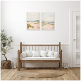 Two-piece framed canvas wall art depicting soft, pastel-colored landscapes with sandy dunes and distant water under a pale sky, hung above a wooden bench with white cushions.