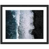 Aerial view of white ocean waves crashing against a textured black volcanic sand beach in Iceland, framed in black with a white mat border.