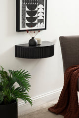 Black semi-circular wall-mounted side table with vertical ribbed texture, decorated with a black vase holding red berries and a small light cup, above a leafy plant beside a gray armchair.