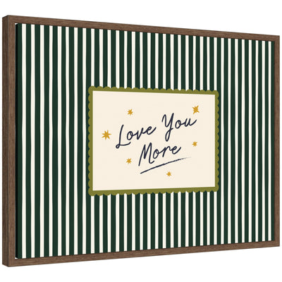 Framed canvas wall art with 'Love You More' quote in striped green background, displayed above bed in a cozy room.