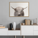 Framed print of a Highland cow with long beige fur and wide curved horns centered on a light background, hanging above a modern white and wood cabinet with vases and books.