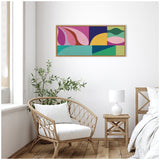 Rectangular abstract canvas with colorful geometric shapes in pink, purple, yellow, green, and teal, framed and hung above a wicker chair in a bright room.