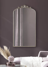 Arched wall mirror with ornate silver detailing on top, reflecting a curtain, mounted on a deep mauve wall above a lavender chaise lounge and a purple glass vase with pampas grass and flowers.