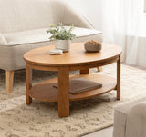 Oval wooden coffee table with a lower shelf, placed on a textured beige rug between beige upholstered seating, decorated with a small potted plant and bowl on top.