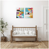Two framed abstract canvas artworks with colorful geometric shapes and patterns hung side by side above a wooden bench with white cushions in a minimalist room.