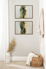 Two framed abstract canvas artworks with vertical, blurry green, beige, and black shapes hanging vertically on a pale wall above a wicker basket with white fabric and a white vase holding pampas grass.