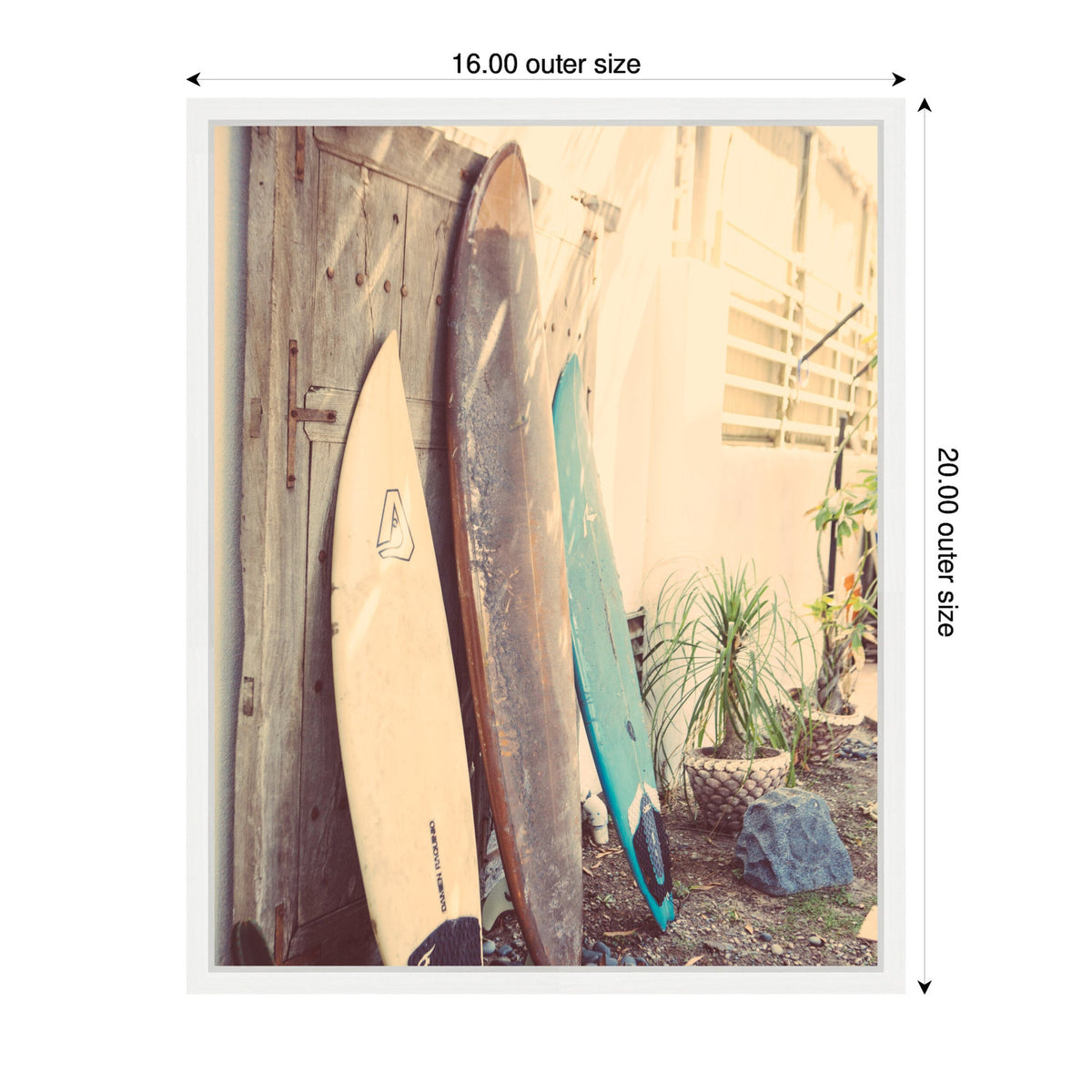Three vintage surfboards in beige, brown, and teal leaning against an aged wooden door in a sunlit outdoor setting with potted plants and gravel ground.