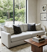 Light gray three-seat sofa with two black patterned and two light parchment pillows, positioned by large window overlooking a green garden, beside a wooden coffee table with books and decor.