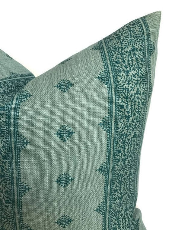 Teal blue pillow cover with a subtle woven texture, featuring small geometric diamond motifs and a detailed floral border pattern along the edges.