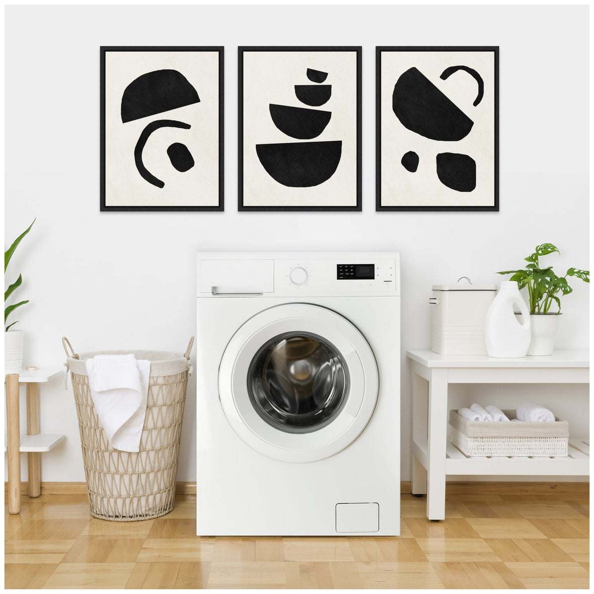 Three black geometric abstract shapes on white background in black frames above a white front-loading washing machine in a bright laundry room with wicker basket and white shelving.