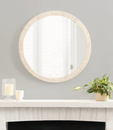 Round wall mirror with a textured light wood frame hanging above a white mantel decorated with a plant in a beige pot and two candles on glass holders.