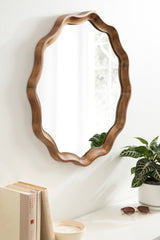Round wall mirror with a scalloped wooden frame in natural brown tones, reflecting a window and hung on a white wall above a table with books, sunglasses, and a potted plant.