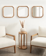 Three square wooden-framed mirrors with rounded corners arranged horizontally above a round wooden side table holding a beige vase with pampas grass, flanked by two cream-colored armchairs with wooden arms.
