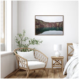 Framed canvas print of a serene green lake surrounded by rocky hills, displayed above a wicker chair and wooden side table with a white lamp in a bright, minimalist room.
