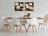 Two framed floral paintings of white peonies with dark green leaves on a black background, hung side by side above a modern dining table with white chairs and a wooden chair.