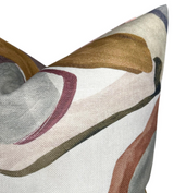 Close-up of a fabric pillow cover showing abstract shapes in muted gold, gray, pink, and brown on a pale beige background with a textured weave.