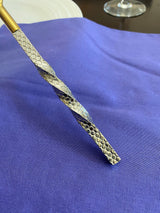 Close-up of a twisted, textured stainless steel utensil handle with a hammered pattern, resting on a blue fabric surface with a white plate and wine glass in the background.