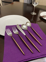 Set of five hammered stainless steel utensils with brown handles including two forks, knife, and two spoons, laid on a purple cloth napkin with white plate and wine glass nearby