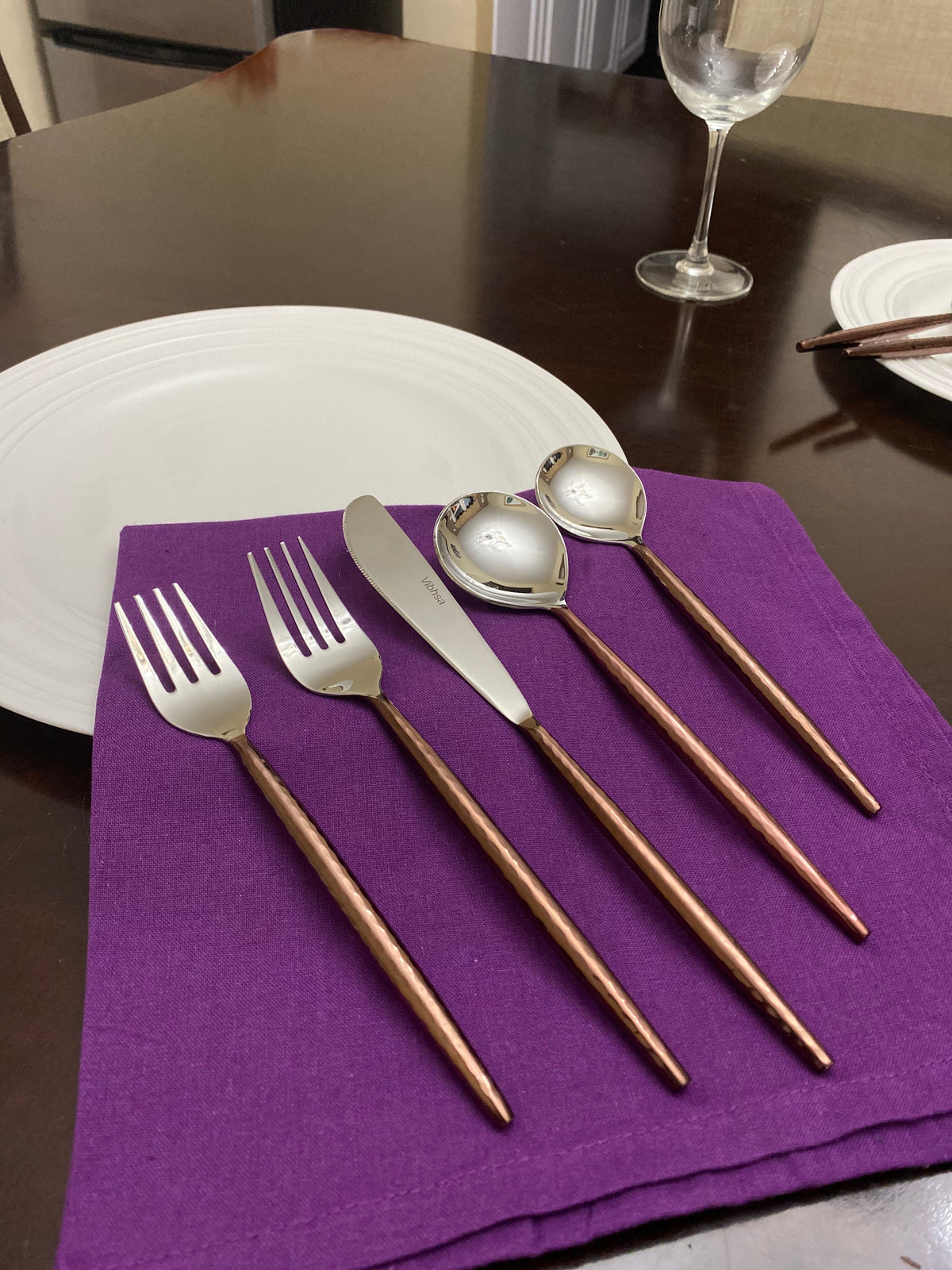 Set of five hammered stainless steel utensils with brown handles including two forks, knife, and two spoons, laid on a purple cloth napkin with white plate and wine glass nearby