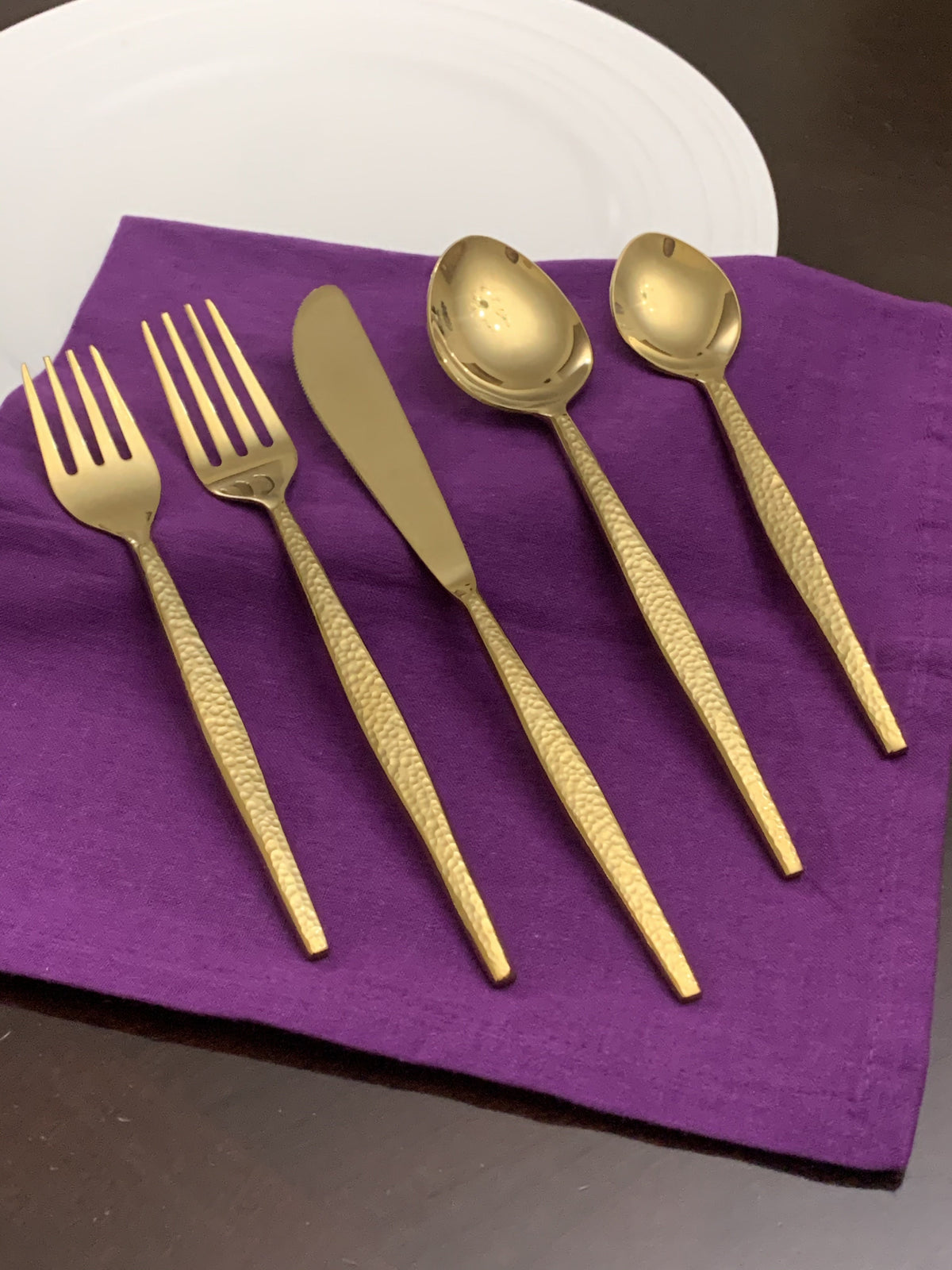 Five hammered gold flatware pieces including two forks, a butter knife, and two spoons arranged on a folded purple cloth with a white plate edge visible.