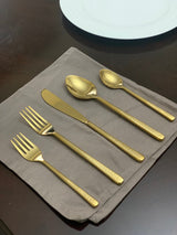 Set of five gold flatware pieces including two forks, a knife, and two spoons with textured handles displayed on a gray cloth napkin with a white plate edge visible above