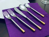 Five-piece silver flatware set including two forks, a knife, and two spoons, each with gold-colored tips, laid out on a purple cloth beside a white plate.