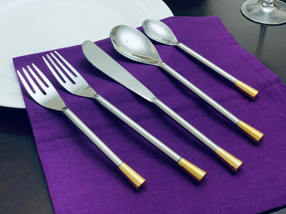 Five-piece silver flatware set including two forks, a knife, and two spoons, each with gold-colored tips, laid out on a purple cloth beside a white plate.