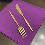 Hammered gold butter knife and four-tine fork with sleek handles placed on a purple cloth on a dark surface