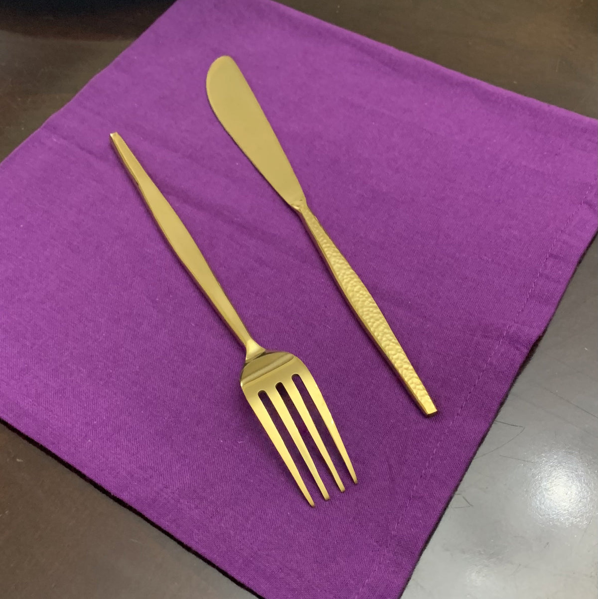 Hammered gold butter knife and four-tine fork with sleek handles placed on a purple cloth on a dark surface