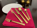Five-piece gold flatware set with textured Egyptian-inspired pattern, including two forks and three spoons, displayed on a bright pink cloth beside a white plate.