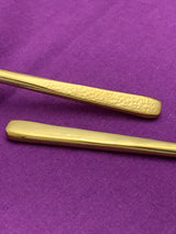 Close-up of two gold flatware handles, one with a smooth finish and the other featuring a textured hammered pattern, against a purple fabric background.