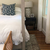 A bedroom corner with a white bed featuring a navy-blue floral-patterned pillow, a dark wooden nightstand with a beige patterned lamp, and white curtains framing a window.