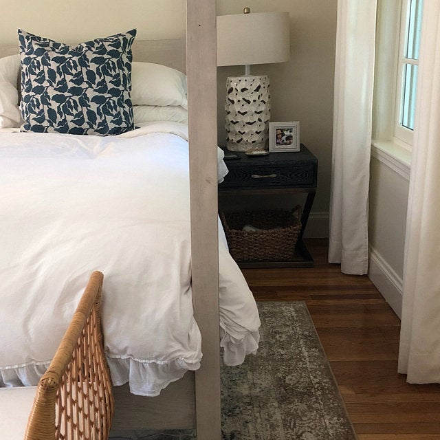 A bedroom corner with a white bed featuring a navy-blue floral-patterned pillow, a dark wooden nightstand with a beige patterned lamp, and white curtains framing a window.