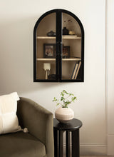 Black-framed wall cabinet with arched top and glass doors, containing decor items, mounted above a black round side table with a plant and next to an olive-green armchair.