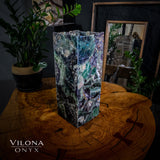 Tall rectangular fluorite lamp with multicolored purple, green, and blue hues on a polished wooden table with circular grain and butterfly joints, surrounded by plants and a mirror.