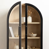 Tall arched wall cabinet with black frame and glass doors revealing light wood interior shelves decorated with books, ceramics, and a vase with dried flowers.