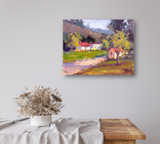 Impressionist-style landscape painting with a red-roofed house, green fields, a tree, and distant hills, mounted on a light gray wall above a wooden table with a white cloth and dried flowers.