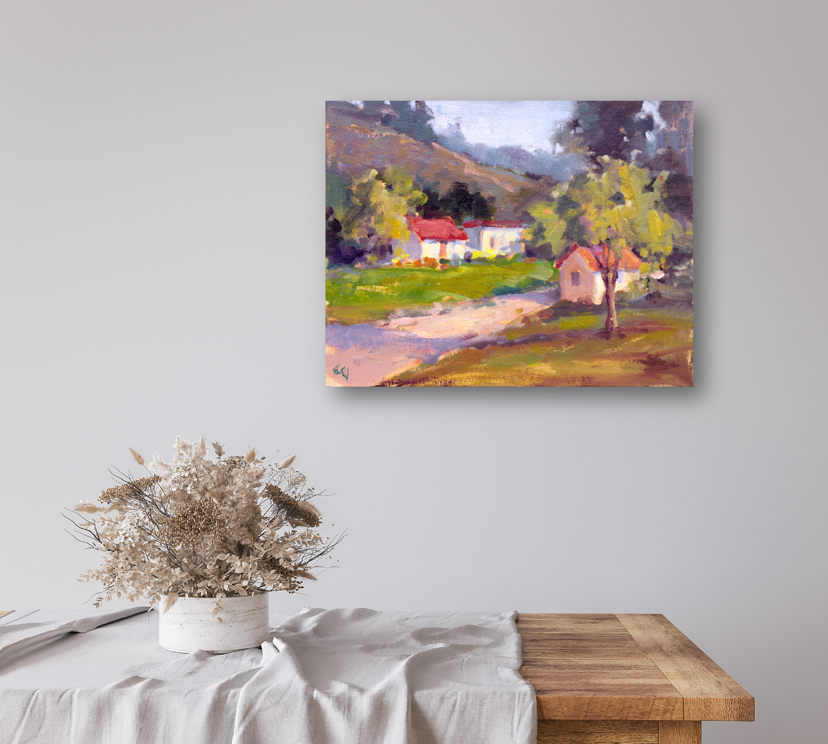 Impressionist-style landscape painting with a red-roofed house, green fields, a tree, and distant hills, mounted on a light gray wall above a wooden table with a white cloth and dried flowers.