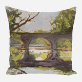 Square pillow featuring painted scene of a stone bridge with three arches over reflective water, surrounded by green trees and branches against a cloudy sky.