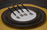 Four small cocktail forks with twisted black handles and silver prongs arranged on concentric white and textured dark plates over a yellow woven placemat.
