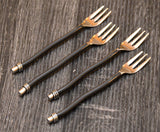 Four cocktail forks with shiny gold-toned heads and black twisted handles, each ending with silver rings, arranged diagonally on a dark textured surface.