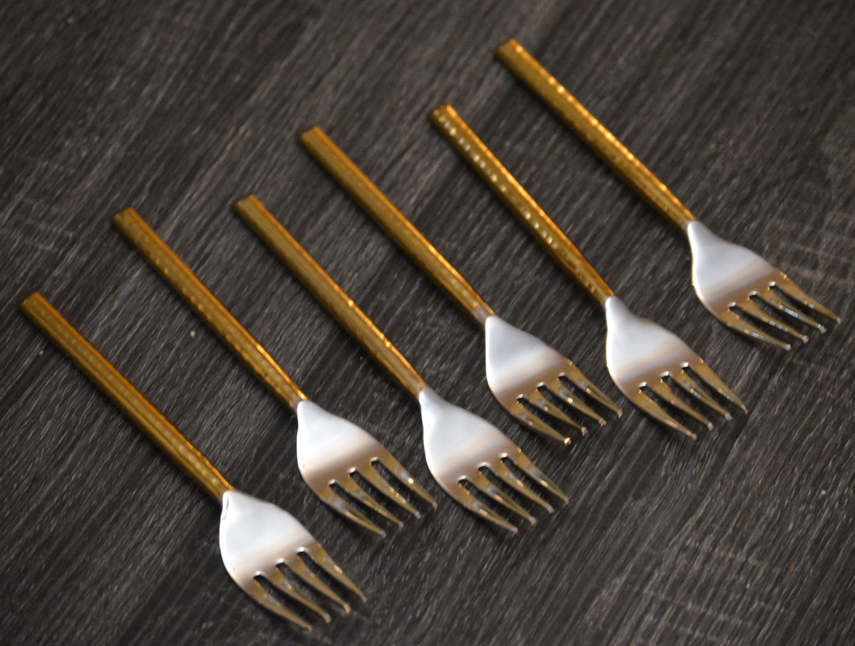 Six dinner forks with golden metal handles and silver tines arranged diagonally on a dark textured surface.
