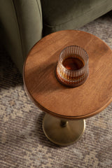 Round wood top side table with a brass pedestal base, holding a textured glass tumbler with amber liquid, positioned near olive green furniture on a patterned rug.