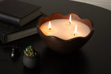 Dark brown metal candle bowl with three lit wicks and a wavy rim, placed on a black round table beside a small black pot of matches and stacked books.