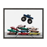 Stacked vintage Matchbox cars in various colors beneath a blue monster truck toy balanced in mid-air, set against a plain white background with wood grain floor.