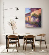Dining area with four wooden chairs around a white table, featuring a colorful impressionist canvas print of a sunlit house with greenery on the wall above.