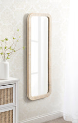 Tall rectangular wall mirror with rounded corners and a wide, light wood grain frame hanging on a patterned cream wall above white furniture with woven drawer fronts.
