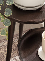 Close-up of dark brown textured three-legged plant stands with scalloped edges, holding white ceramic pots and accompanied by green round-leafed plant stems.