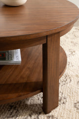 Close-up of a round wood coffee table with a smooth walnut finish, featuring a thick tabletop, sturdy vertical legs, and a lower shelf holding books.